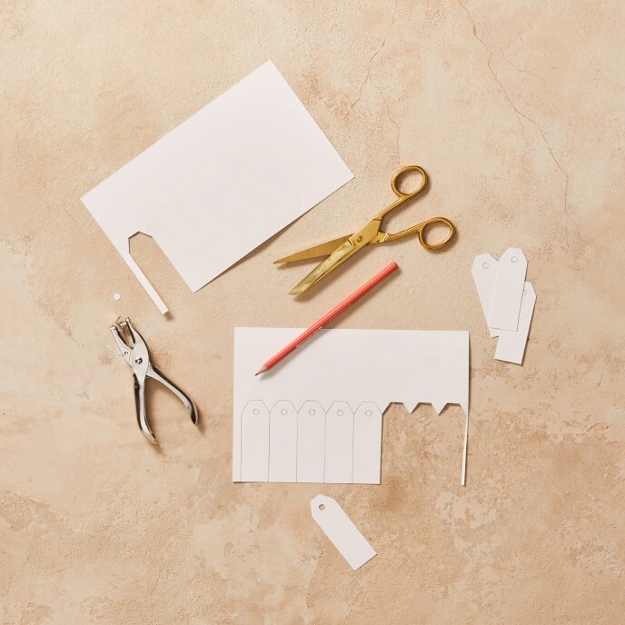 Step 1 of the DIY project: A close-up of a hand cutting out a rectangular piece of cardstock with a patterned design to create the base for the Christmas place card.