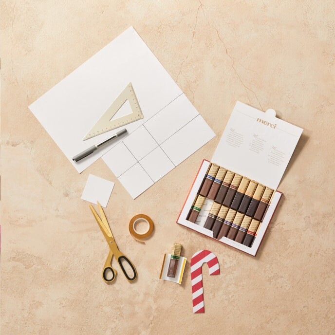 Étape 3 : Une main fixant un support en papier blanc plié au dos d’une étiquette-cadeau en forme de canne en bonbon à l’aide d’un ruban adhésif double face, le préparant à contenir du chocolat merci.