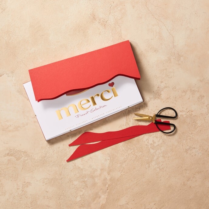 Step 3: An image showing a card made from red and pink rectangles glued together folded over a merci package, with a decorative shape drawn and partially cut out to ensure the "merci" lettering remains visible.